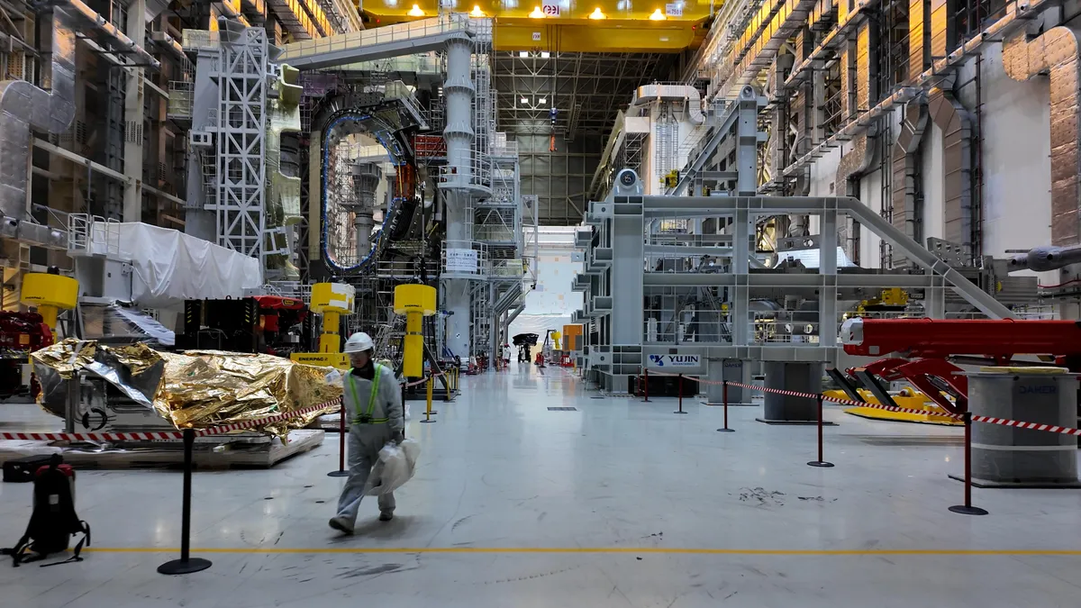 Внутри сборочного цеха токамака компании ITER – высокое здание, заполненное компонентами токамака, и проходящий мимо рабочий.