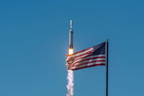Искусственный интеллект SpaceX: что способствует звездам, а что угрожает НАСА? 1 Ракета SpaceX Falcon Heavy стартует в ясное голубое небо с развевающимся американским флагом на переднем плане.