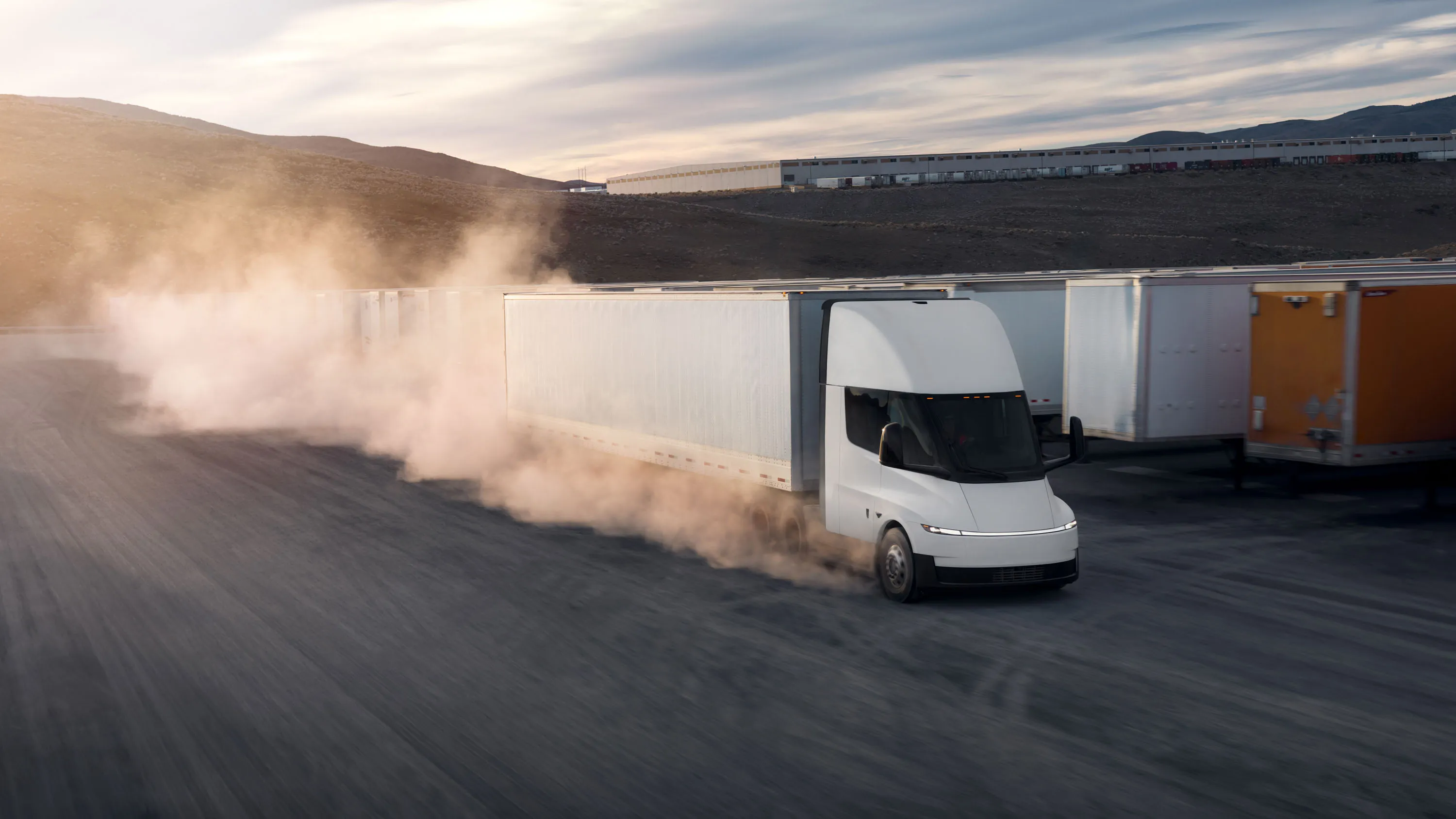 Грузовик Tesla поднимает пыль, двигаясь в районе завода и рядов прицепов.