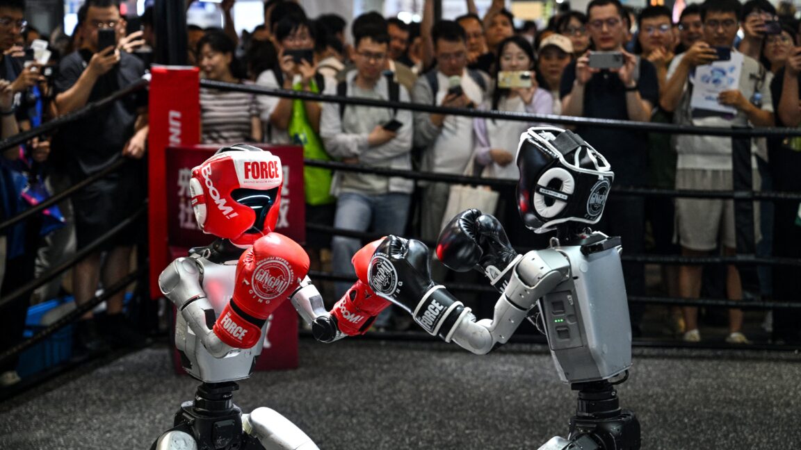 Гуманоидные роботы, управляемые дистанционно компанией Unitree Robotics, принимают участие в поединке во время Всемирной конференции по искусственному интеллекту (WAIC) в Шанхае 18 июля 2026 года. На переднем плане слева изображен один робот в красном шлеме и боксерских перчатках, а справа — робот в черной экипировке. На заднем плане — толпа зрителей, стоящих за канатами боксерского ринга.