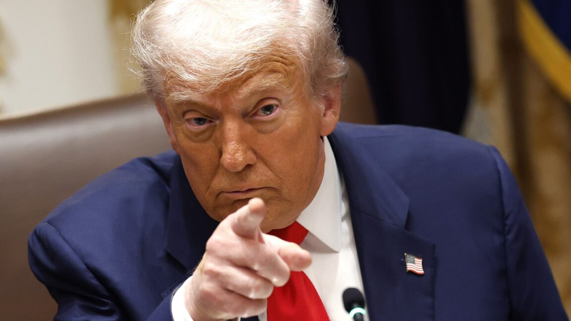 Президент Дональд Трамп сидит на стуле и указывает указательным пальцем правой руки во время совещания.