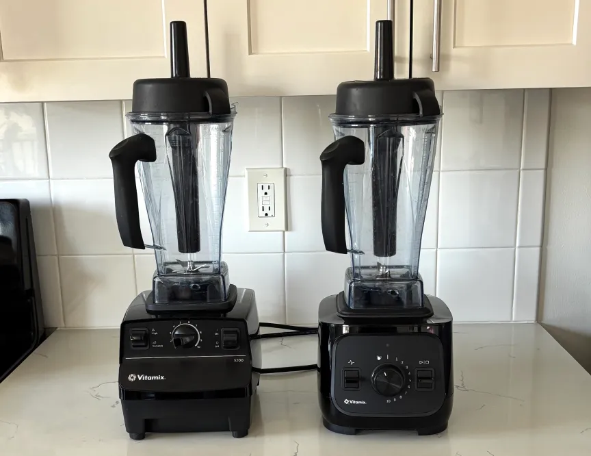 Слева — блендер Vitamix 5200, справа — VX1, оба черного цвета, на белой мраморной кухонной столешнице.