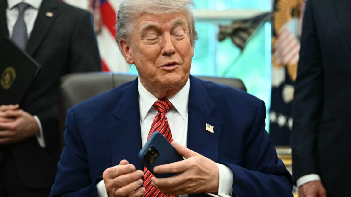 Президент Трамп сидит за своим рабочим столом в Белом доме, держа в левой руке смартфон.