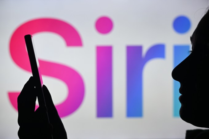 Изображение женщины, держащей мобильный телефон на фоне логотипа Siri, отображаемого на экране компьютера.